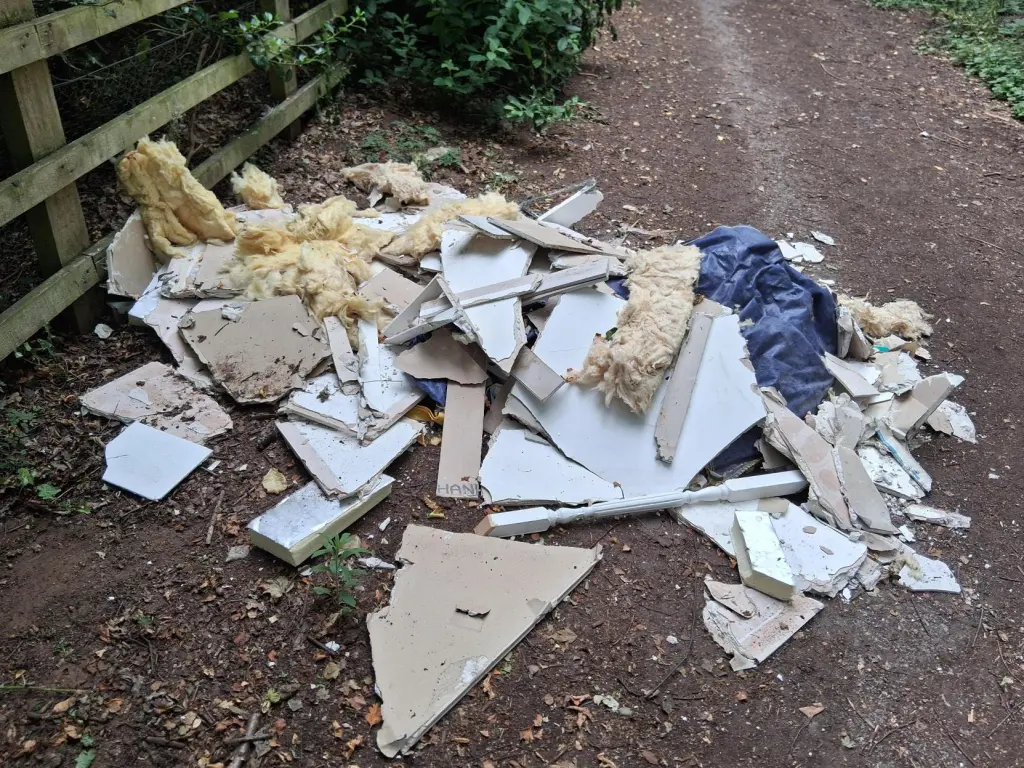 Broken plasterboard, insulation and carpet dumped across a dirt track beside a fence