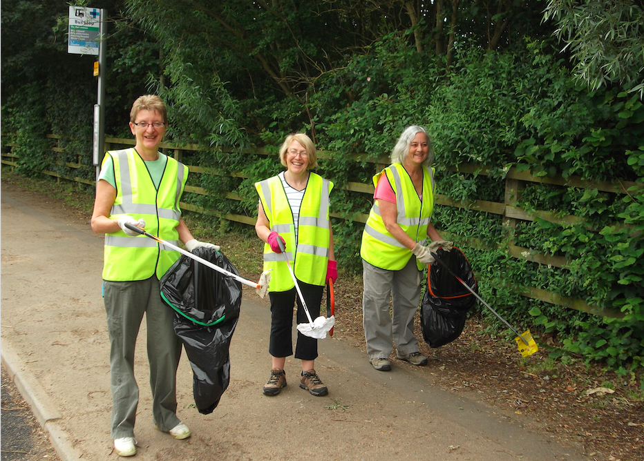 Three volunteers in hi-vis vests holding litter pickers and bin bags beside a path