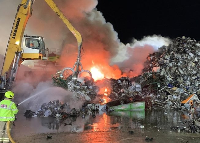 Firefighter spraying water on a burning scrap-metal pile beside a material handler