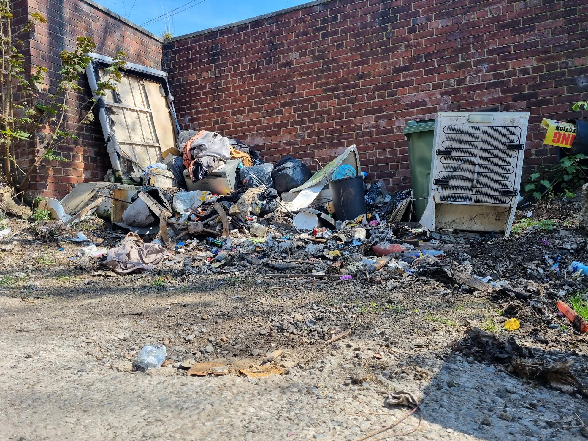 Large fly-tipped pile of bags, a mattress and debris against a brick wall in a yard