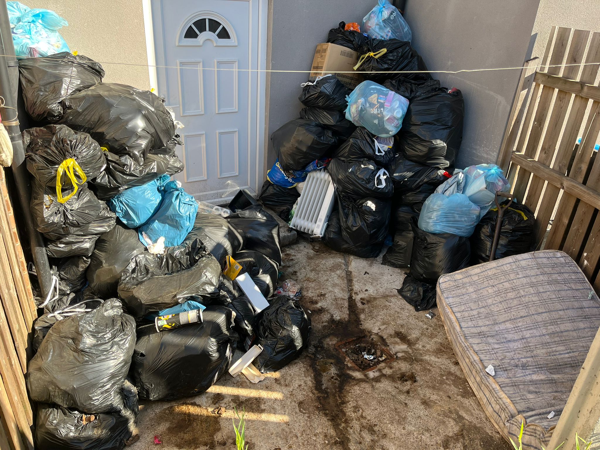 Bin bags, a mattress and household waste piled in a yard beside a back door