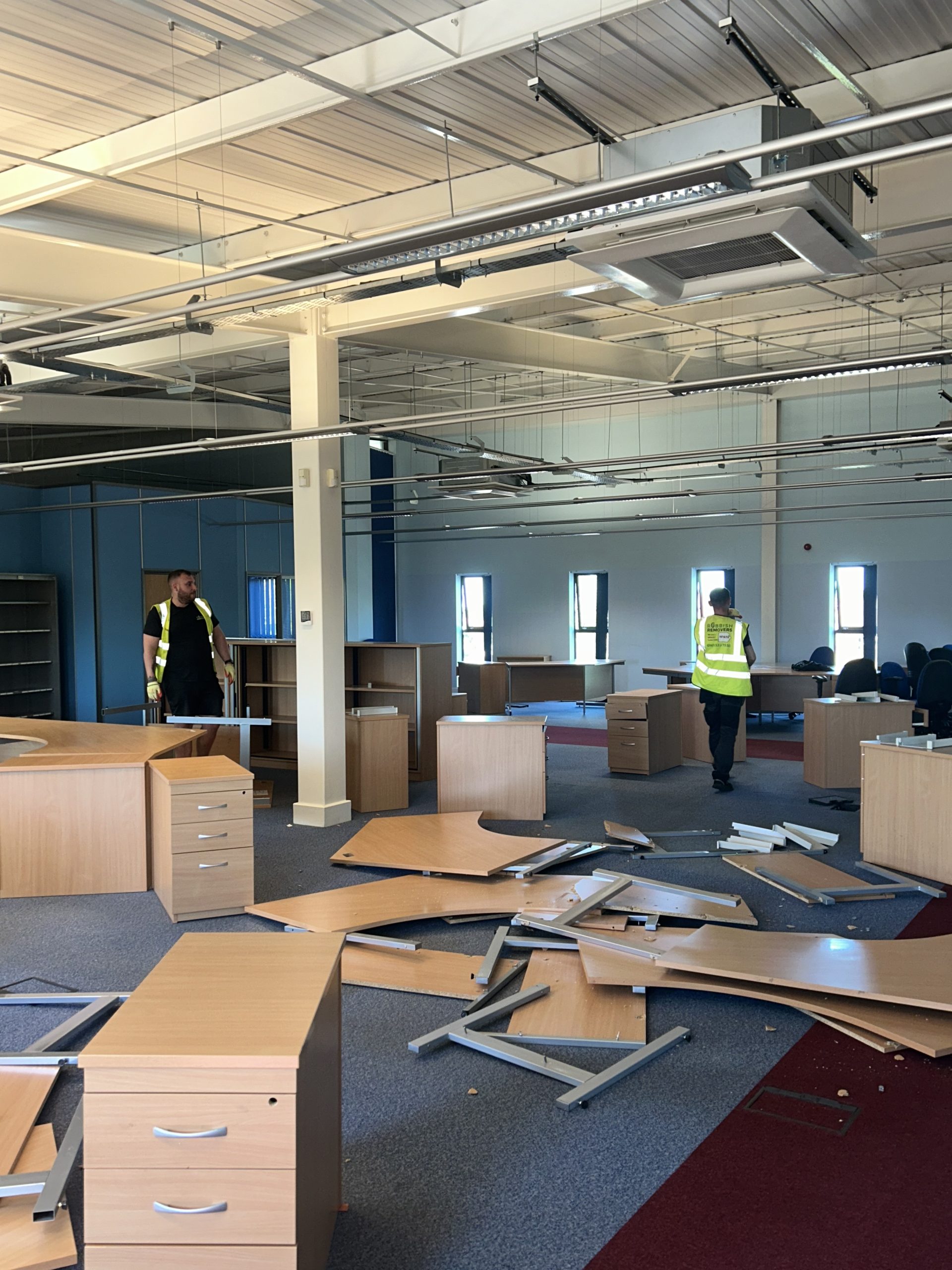 Large open-plan office with dismantled wooden desks and pedestals on the floor, and a worker in hi-vis