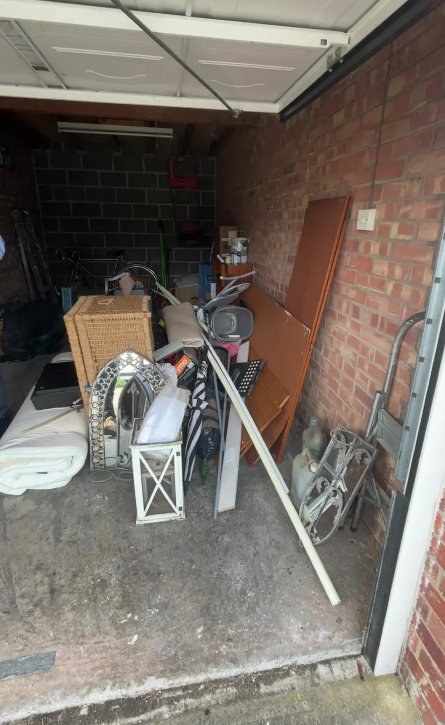 Cluttered garage with its door open, filled with furniture, boards and household items