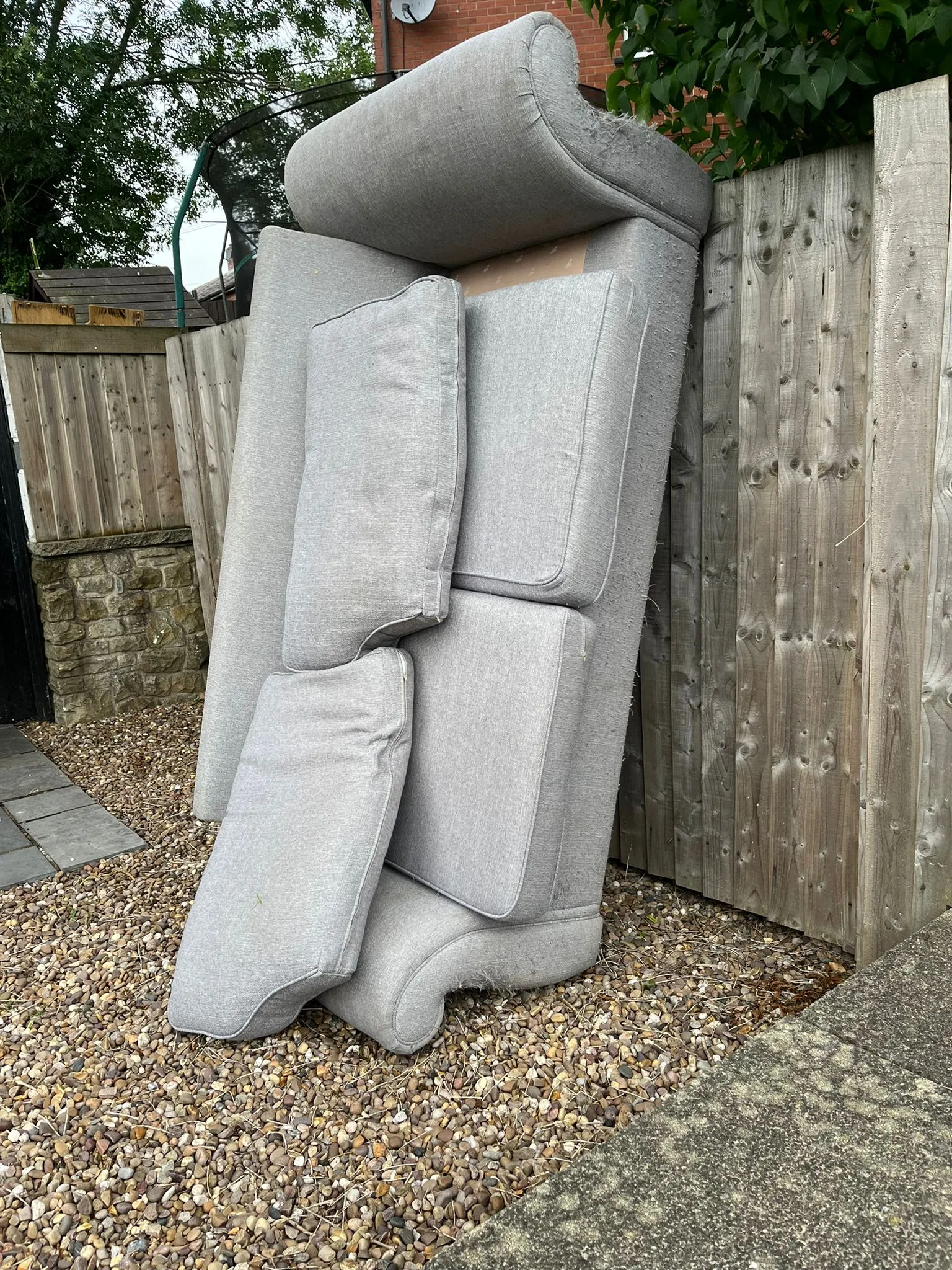 Old grey sofa and cushions dumped against a garden fence
