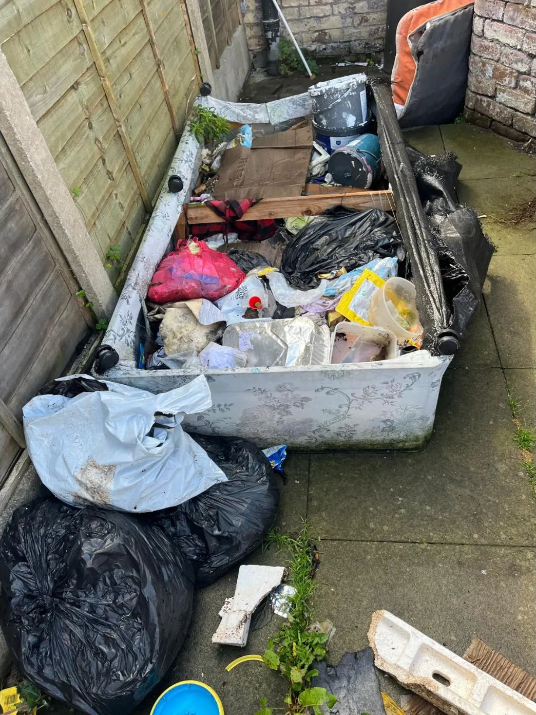 Old bathtub filled with rubbish, with bin bags beside it along a fenced side path