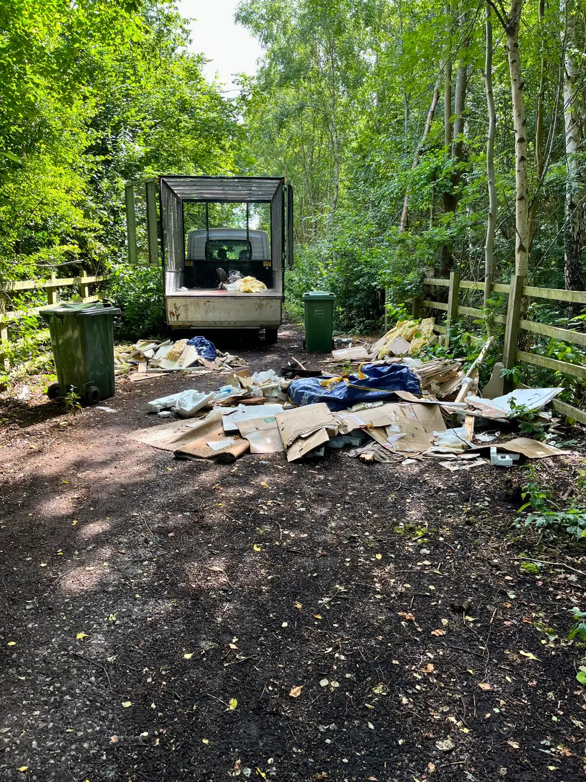 Fly-tipped rubbish and cardboard scattered along a woodland track, with a caged tipper truck behind