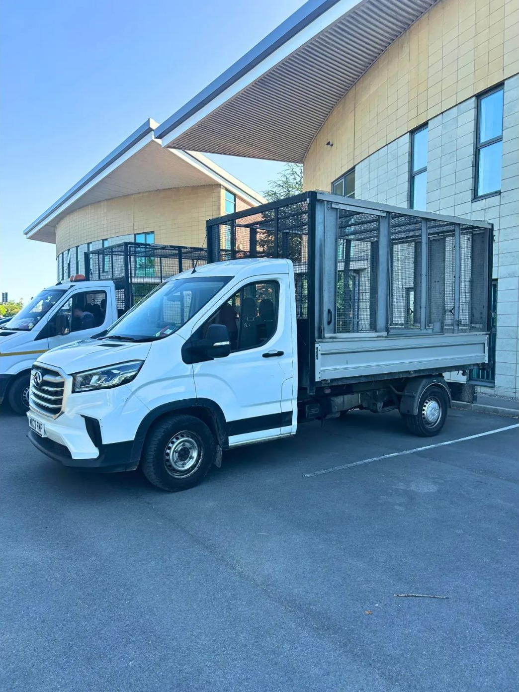 Two white caged tipper trucks parked outside a modern office building
