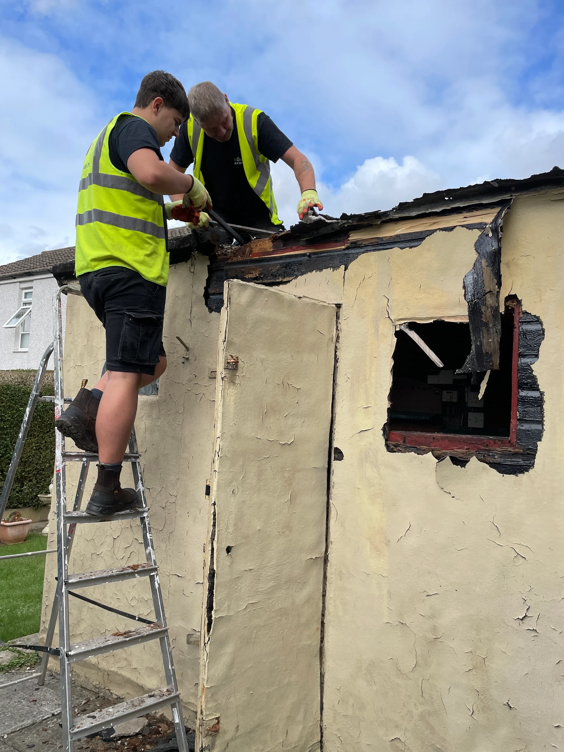 Two workers in hi-vis on a ladder stripping the roof from a timber shed