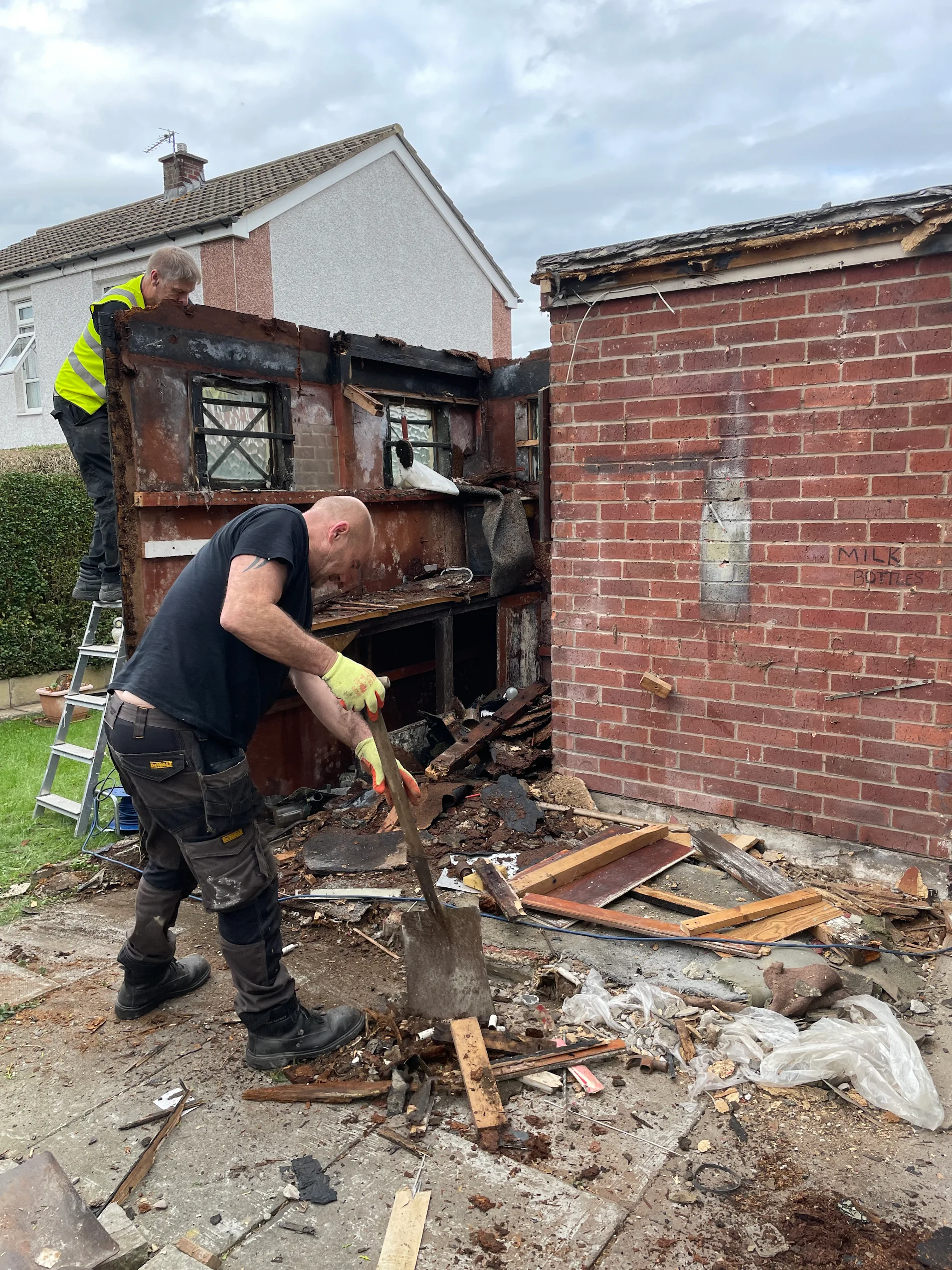 Workers dismantling a timber shed beside a brick outhouse, with debris on the ground