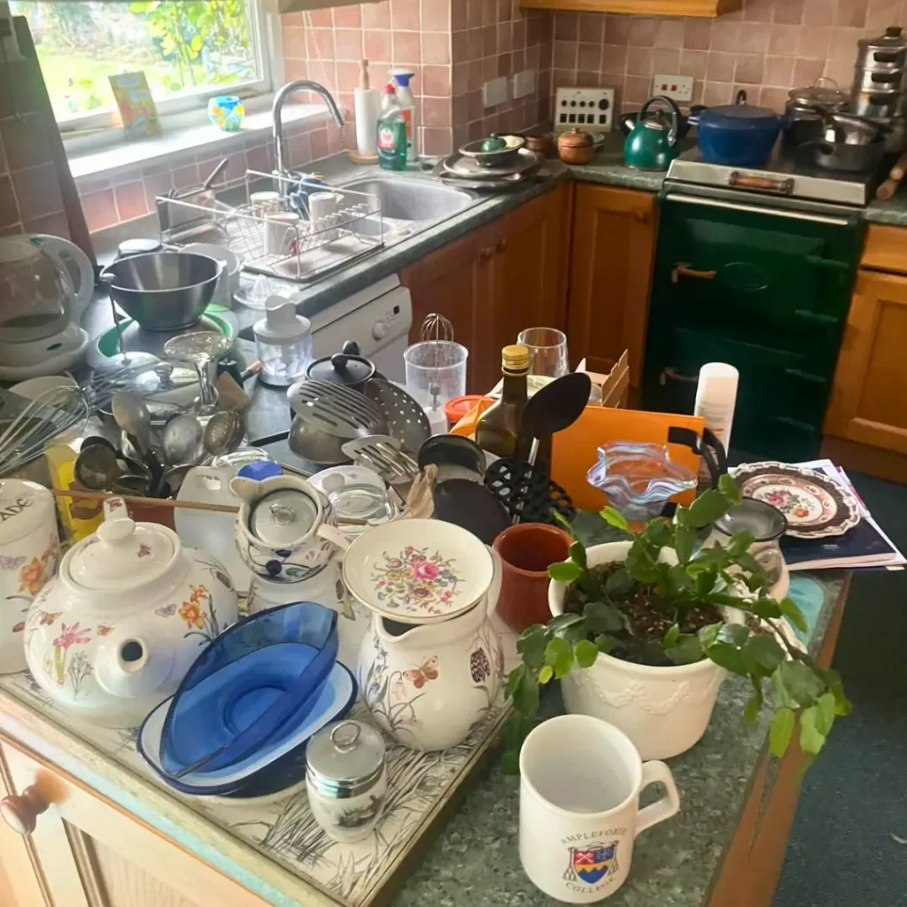 Kitchen before a house clearance with cluttered worktops, dishes and household items ready for removal.